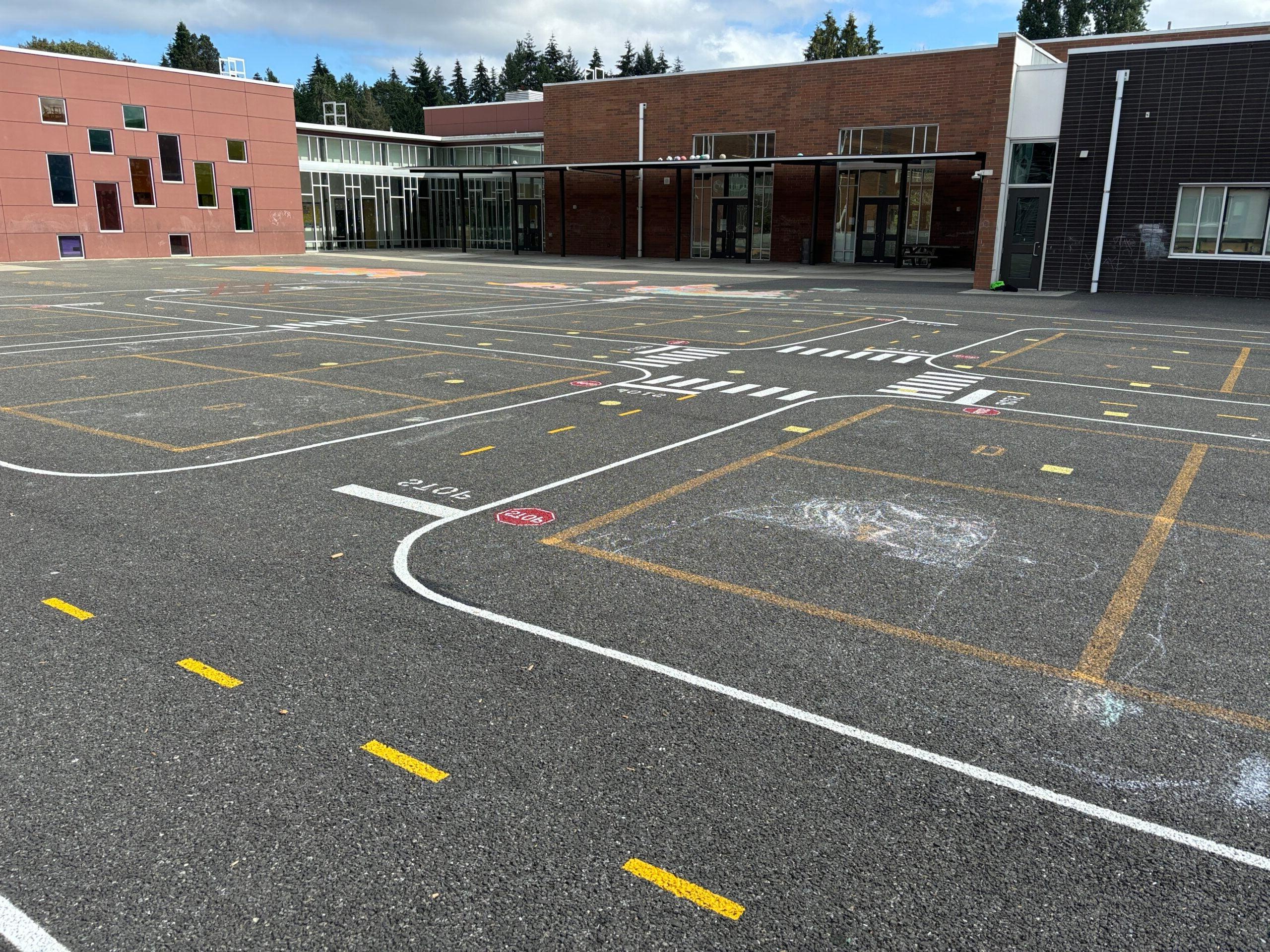 Miniature painted road curves around four square court and student chalk art. On black top with school in background.